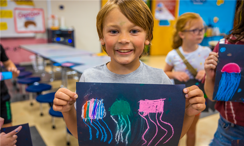 A boy with dark blond hair holds up a piece of art